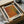 May include: Framed vintage-style art print with geometric orange, yellow, and gray halftone dot patterns forming angular shapes on cream background, displayed in black frame within cardboard shipping box surrounded by protective tissue paper and wooden dowels on table surface.