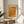 May include: Framed art print with orange dotted pattern and Japanese text displayed on wooden round side table next to beige upholstered chair. Natural sunlight streams across white walls creating soft shadows. Green plant leaves visible in corner. Warm minimalist interior styling with neutral tones and natural wood frame.