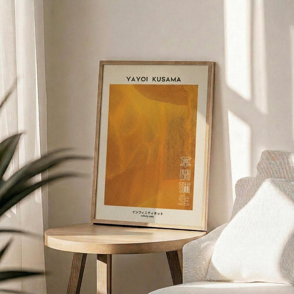 May include: Framed art print with orange dotted pattern and Japanese text displayed on wooden round side table next to beige upholstered chair. Natural sunlight streams across white walls creating soft shadows. Green plant leaves visible in corner. Warm minimalist interior styling with neutral tones and natural wood frame.