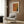 May include: Framed poster with orange, yellow and teal geometric abstract design displayed on wood paneled wall above modern beige armchair with patterned throw pillow. Room features brass floor lamp, dark round side table with open book, and sheer curtains. Poster shows dotted halftone pattern creating angular shapes in warm autumn colors within light wood frame.