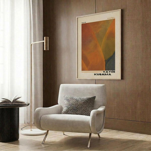 May include: Framed poster with orange, yellow and teal geometric abstract design displayed on wood paneled wall above modern beige armchair with patterned throw pillow. Room features brass floor lamp, dark round side table with open book, and sheer curtains. Poster shows dotted halftone pattern creating angular shapes in warm autumn colors within light wood frame.