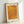 May include: Framed poster print leaning against white wall showing orange and yellow dotted pattern artwork with flowing curved lines made of small dots. Light wood frame surrounds cream-colored mat with black text at top and bottom including Japanese characters. Soft natural lighting creates gentle shadows on textured wall surface.