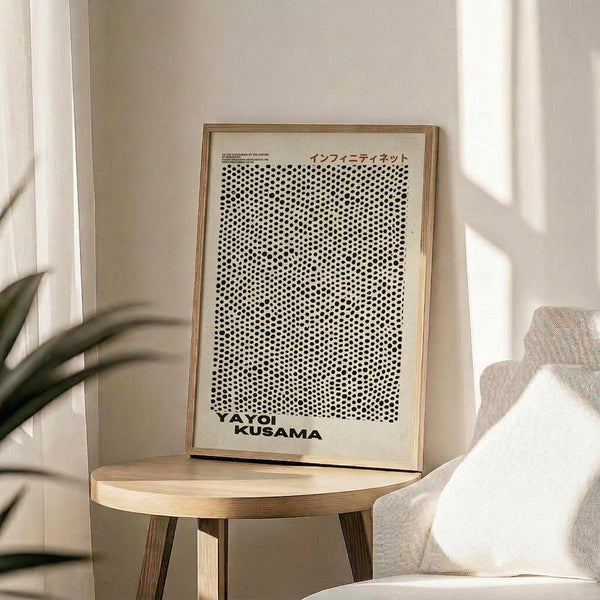 May include: A framed art print featuring black polka dots densely arranged on white background with Japanese text and "YAYOI KUSAMA" at bottom, displayed on a light wood round side table next to a beige upholstered chair with white pillow. Natural sunlight streams across the neutral-toned room with white walls and a green plant visible in corner.