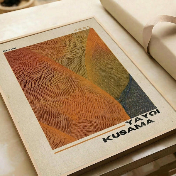 Abstract geometric artwork featuring overlapping curved forms in warm orange, yellow, and green tones with halftone dot pattern texture. The composition shows flowing, organic shapes against a cream-colored background. Text reading "YAYOI KUSAMA" appears at bottom right, with "Tokyo 1998" in upper left corner. The print is displayed on white paper alongside rolled paper tubes on a neutral surface.