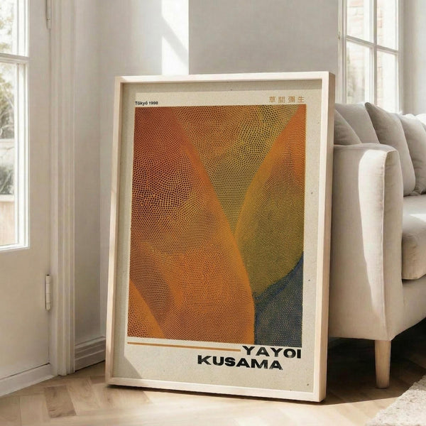 May include: Framed poster with orange, yellow, and teal dotted pattern artwork leaning against white wall next to beige upholstered chair. The print features halftone dots creating flowing, wave-like forms in warm sunset colors transitioning to cool blue-green. Text reads "YAYOI KUSAMA" and "Tokyo 1999" on cream-colored paper within light wood frame. Bright natural light streams through tall windows onto hardwood floors.