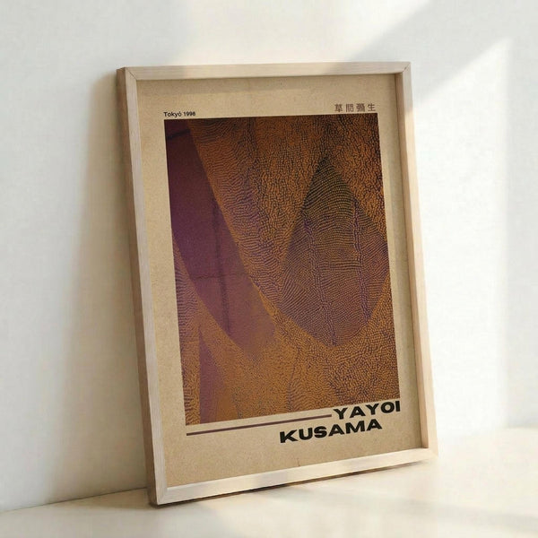A framed art print leaning against a white wall, featuring an abstract composition in warm brown and burgundy tones with intricate dotted patterns and flowing organic shapes. The artwork has a textured, stippled appearance with curved forms that create depth and movement. Text reading "YAYOI KUSAMA" appears at the bottom, with "Tokyo 1998" in the upper left corner. The print is displayed in a simple light wood frame on a cream-colored mount.