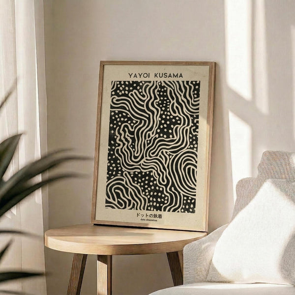 May include: Black and white abstract art print with flowing wavy lines and scattered dots pattern, displayed in natural wood frame on round wooden side table next to beige upholstered chair. Sunlight streams across white wall creating geometric shadows, with green plant leaves visible in corner of bright, minimalist room setting.