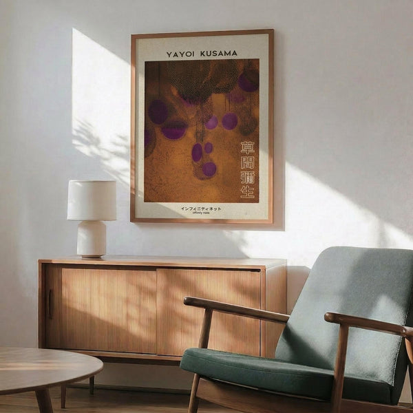May include: Yayoi Kusama exhibition poster featuring abstract dotted pattern in warm orange and brown tones with purple circular forms, displayed in thin wooden frame above mid-century modern walnut credenza. Scene includes sage green upholstered armchair with wooden frame, white ceramic table lamp, and natural sunlight casting shadows on white wall.