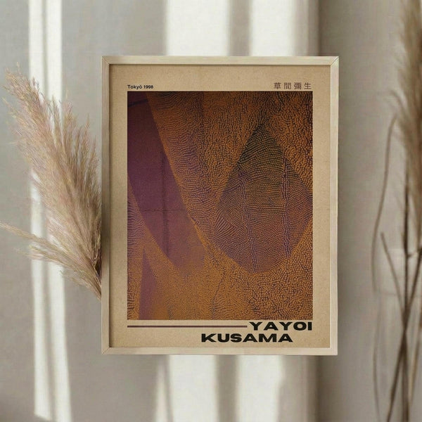 May include: Framed art print with warm orange and brown abstract textural pattern, resembling organic or cellular forms with deep burgundy accents. Artist name displayed at bottom in bold black text. Light wood frame mounted on neutral wall beside dried pampas grass stems, creating soft shadows in natural lighting.