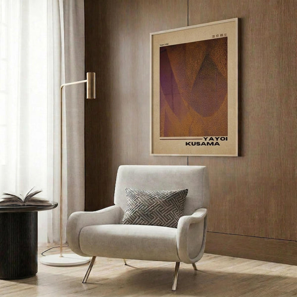 May include: Modern interior with cream upholstered armchair featuring geometric patterned pillow, gold floor lamp, dark round side table with open book, and framed art poster on wood-paneled wall. Poster shows abstract brown and purple curved forms with dotted texture in cream matting. Warm neutral tones throughout scene with natural lighting from window.