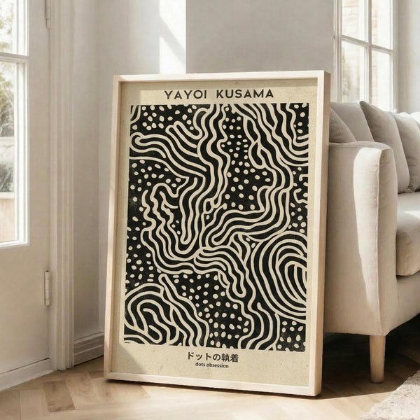 May include: Framed black and white abstract art print leaning against white wall in bright living room. The artwork features flowing wavy lines and scattered dots in Kusama's signature style, with "YAYOI KUSAMA" text at top and Japanese characters below. Light wood frame, positioned next to beige upholstered chair near large windows with natural light streaming in.