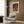 May include: Modern reading corner with beige upholstered armchair and geometric patterned pillow, featuring framed art poster with orange and brown background, purple polka dots, and Japanese text. Wood paneled wall backdrop, brass floor lamp, dark round side table with open book, and white curtains visible.