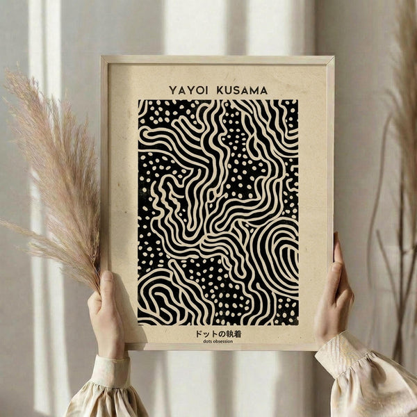 May include: Person holding a framed black and white abstract art print featuring flowing wavy lines and polka dots in Yayoi Kusama's distinctive style. The artwork is mounted on cream paper within a light wood frame. One hand also holds dried wheat or pampas grass stems. Soft natural lighting creates a minimalist aesthetic against neutral curtained background.