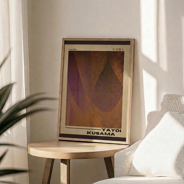 May include: Framed art print with warm orange and purple abstract leaf-like forms displayed on a light wood round side table next to a beige upholstered chair. Natural sunlight streams across white walls, with a plant's green leaves visible in the corner, creating a serene modern living room setting.