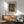 May include: Black and white abstract art print with flowing wavy lines and dotted patterns in wooden frame, displayed above mid-century modern walnut credenza with white ceramic table lamp. Sage green upholstered armchair with wooden frame sits nearby on wooden floor. Soft natural light creates shadows on white wall in minimalist living room setting.