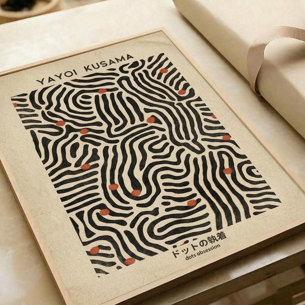 Minimalist art print featuring flowing black curved lines creating an abstract maze-like pattern on cream background, with small orange-red dots scattered throughout. The print sits in a wooden frame on white paper against neutral fabric surfaces, creating a warm, modern aesthetic with organic flowing forms.