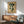 May include: Framed botanical poster with dark green leaf forms on warm beige background, displayed above wooden sideboard with table lamp in modern living room setting. Mid-century style chair with sage green upholstery sits nearby. Natural sunlight creates geometric shadows on white wall.