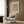 May include: Contemporary interior with beige upholstered armchair featuring geometric patterned pillow, gold floor lamp with white marble base, dark round side table with open book, and framed art print on wood-paneled wall. The artwork displays flowing black linear patterns resembling topographical waves on cream background in light wooden frame. Neutral color palette with warm wood tones and soft lighting from window with curtains.