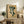 May include: Framed botanical poster with dark green leaf shapes arranged vertically on warm beige background, displayed on rustic wood sideboard alongside fern plant, stacked books, and ceramic vase in neutral-toned living room setting with upholstered chair visible.