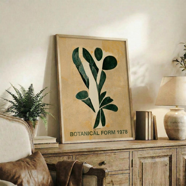 May include: Framed botanical poster with dark green leaf shapes arranged vertically on warm beige background, displayed on rustic wood sideboard alongside fern plant, stacked books, and ceramic vase in neutral-toned living room setting with upholstered chair visible.