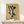 May include: Framed botanical print featuring green leaf shapes arranged vertically on aged beige paper background, displayed on light wood surface against white wall. Small white wrapped gift box with twine sits beside the wooden-framed artwork. Vintage-style poster shows various oval and elongated leaves in dark green tones with text at bottom.