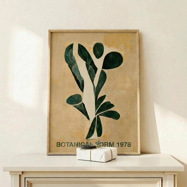 May include: Framed botanical print featuring green leaf shapes arranged vertically on aged beige paper background, displayed on light wood surface against white wall. Small white wrapped gift box with twine sits beside the wooden-framed artwork. Vintage-style poster shows various oval and elongated leaves in dark green tones with text at bottom.
