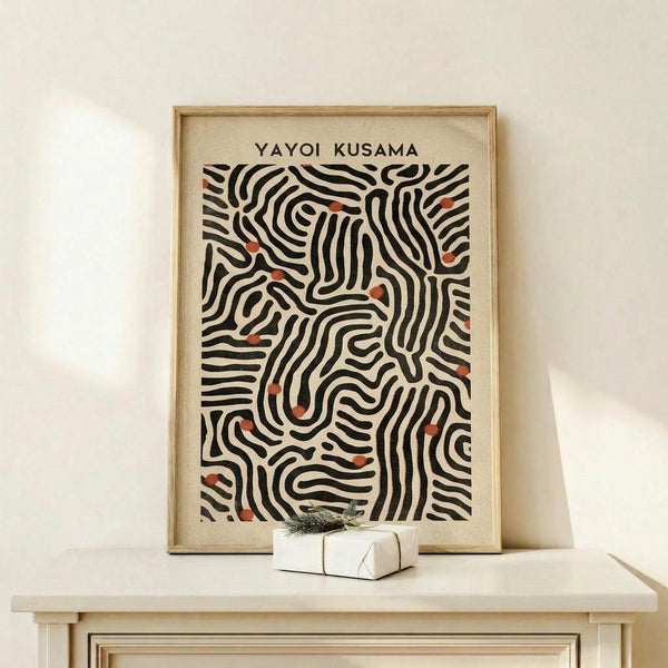 May include: Framed art print featuring abstract black wavy lines with small red-orange dots scattered throughout on cream background, displayed on light wood console table next to small white wrapped gift with evergreen sprig, against white wall with soft natural lighting