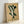 May include: Framed botanical print leaning against white wall corner. Artwork features abstract green leaf shapes arranged vertically on aged cream background with yellow staining. Text reads "BOTANICAL FORM 1978" at bottom. Light wood frame casts subtle shadow on wall. Vintage-inspired minimalist design with organic curved forms.