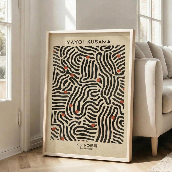 May include: A framed art print leaning against a white wall next to a beige sofa in a bright living room with large windows. The poster features flowing black organic lines creating maze-like patterns with scattered red dots on a cream background. Text reads "YAYOI KUSAMA" at top and Japanese characters at bottom. The print has a light wooden frame and casts a soft shadow on the wooden floor.