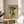 May include: Framed botanical poster with dark green leaves arranged in an artistic spray pattern on cream background, displayed on round wooden side table next to beige upholstered chair. Natural sunlight creates shadows across white wall. Live plant leaves visible in corner of modern, minimalist room setting.