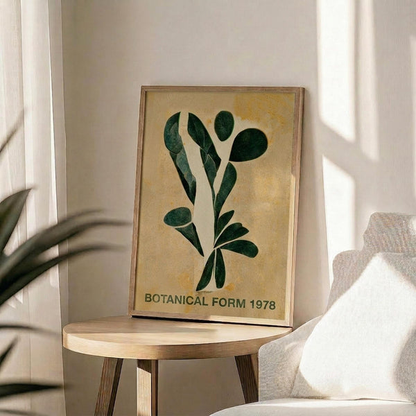 May include: Framed botanical poster with dark green leaves arranged in an artistic spray pattern on cream background, displayed on round wooden side table next to beige upholstered chair. Natural sunlight creates shadows across white wall. Live plant leaves visible in corner of modern, minimalist room setting.
