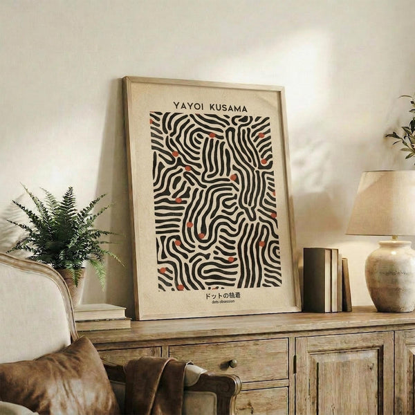 May include: Framed art print featuring black wavy lines with red dots on cream background, displayed on rustic wooden sideboard next to fern plant, ceramic vase, and stacked books in warm-toned living room setting with beige chair visible in foreground