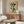 May include: Framed botanical poster with green leaves on warm yellow background mounted above mid-century walnut credenza. Modern living room setting features cream curved chair, round glass coffee table with blue accent, white sculptural lamp, and neutral walls with natural lighting.