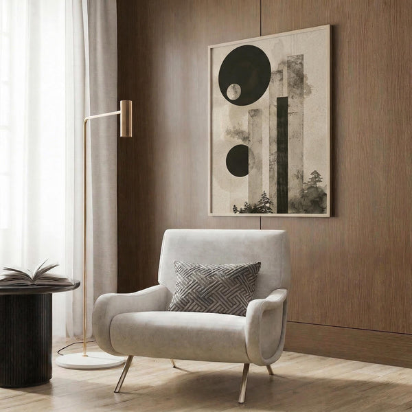 May include: Abstract art print with dark green circles and geometric shapes over textured beige background with watercolor tree silhouettes, displayed in light wood frame on warm wood paneled wall above cream upholstered armchair with geometric pillow, brass floor lamp and side table nearby in modern neutral living room setting