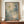 May include: Large framed abstract expressionist artwork with splattered blue, green, and cream paint, white dots, and linear scratches displayed on wooden credenza beside white ceramic vase and stacked books against cream wainscoted wall