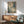 May include: Abstract expressionist artwork in wooden frame hanging above mid-century modern credenza with white table lamp. Features splattered paint in blue, green, beige and white tones with organic flowing forms. Modern living room setting with sage green upholstered chair, round coffee table, and natural sunlight casting shadows on white wall.