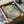Framed abstract expressionist artwork being unpacked from cardboard shipping box with protective wrapping. The piece features soft watercolor-style splatters and brushstrokes in muted blues, greens, and cream tones with delicate line work overlaid throughout. Black frame sits nestled in brown cardboard packaging with white tissue paper and protective corner guards visible around the edges.