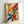 Abstract expressionist artwork with vibrant paint splatters and energetic brushstrokes in yellow, red, blue, green, and black against a cream background. The composition features dynamic drips, splashes, and gestural marks creating movement across the canvas. Displayed in a natural wood frame leaning against a white wall, showing the textured paint application and spontaneous painting technique characteristic of action painting style.