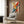 May include: Vibrant abstract expressionist artwork with bold splashes of red, yellow, and blue paint against white background, displayed in thin frame on wood-paneled wall above cream upholstered armchair with geometric patterned pillow, alongside gold floor lamp and dark round side table with open book