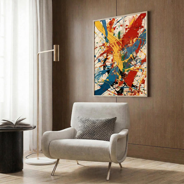 May include: Vibrant abstract expressionist artwork with bold splashes of red, yellow, and blue paint against white background, displayed in thin frame on wood-paneled wall above cream upholstered armchair with geometric patterned pillow, alongside gold floor lamp and dark round side table with open book