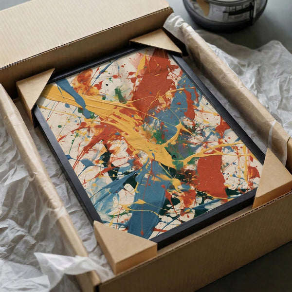 Framed abstract expressionist artwork with vibrant paint splatters in red, blue, yellow, and green against cream background, packed in cardboard shipping box with protective materials and tube packaging visible alongside