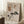 May include: Framed abstract artwork leaning against white wall in sunlit room, featuring beige background with geometric grid, crescent moon, stylized pine tree, and brown rectangular shapes. Natural wood frame, displayed near coffee cup on wooden stool and beige textiles on white fluffy rug.