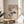 May include: Framed abstract art print displayed on round wooden side table next to beige upholstered chair. Artwork features geometric shapes, crescent moon, and stylized pine tree branch in neutral earth tones. Natural lighting creates soft shadows across cream-colored wall. Green plant leaves visible in corner of minimalist room setting.