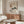 May include: Abstract art print in warm wooden frame hanging above mid-century walnut credenza in minimalist living room. Artwork features geometric rectangles, crescent moon, and stylized pine tree branch in earth tones on textured beige background. Room styled with cream curved chair, glass oval coffee table, white mushroom lamp, and wooden mirror, all in soft natural lighting.