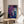 May include: Abstract fluid art print with swirling purple and blue paint creating marble-like patterns with white crackling effects throughout. The artwork is displayed in a thin wooden frame, leaning against a white wall on a round wooden side table. Soft natural lighting creates shadows across the minimalist room setting with a beige upholstered chair and green plant leaves visible in the frame.