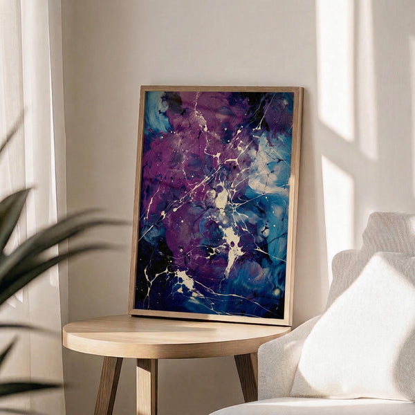 May include: Abstract fluid art print with swirling purple and blue paint creating marble-like patterns with white crackling effects throughout. The artwork is displayed in a thin wooden frame, leaning against a white wall on a round wooden side table. Soft natural lighting creates shadows across the minimalist room setting with a beige upholstered chair and green plant leaves visible in the frame.