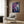 May include: Abstract artwork with purple, blue, and white paint splatters and flowing organic shapes displayed in light wood frame above beige upholstered armchair with geometric patterned pillow. Modern reading corner features brass floor lamp with white marble base and dark round side table with open book. Wood paneled wall and white curtains complete the contemporary interior setting.