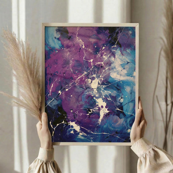 May include: Person in cream sweater holding framed abstract art print with purple, blue, and teal fluid paint splatters and white crackling lines, displayed near dried pampas grass against soft curtained window background