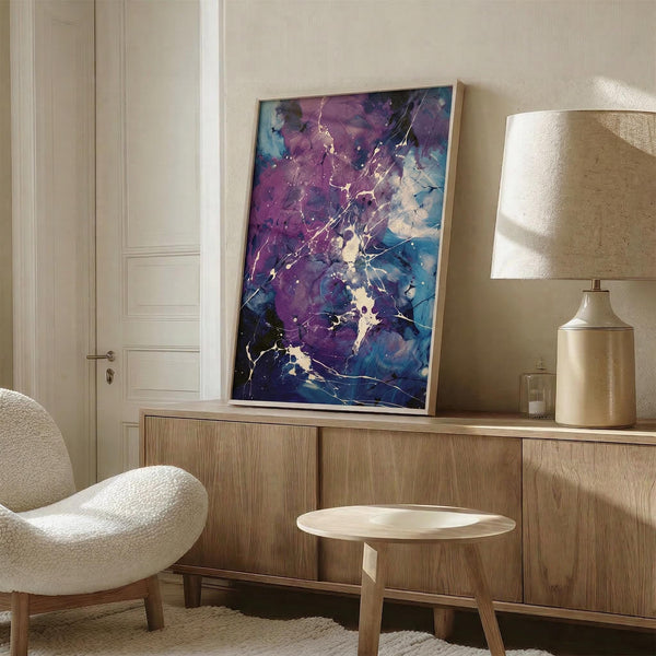 May include: Abstract artwork with purple and blue splatter design featuring gold veining and organic flowing shapes, displayed in thin light wood frame on wooden credenza beside cream table lamp and white upholstered chair in neutral-toned room setting.