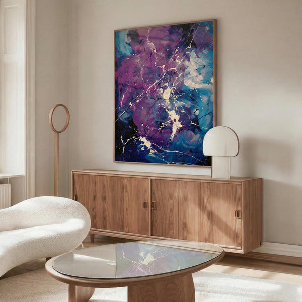May include: Abstract splatter art print with vibrant purple and blue fluid paint effects, white crackling details, and gold metallic accents, displayed in thin gold frame above walnut credenza. Modern living room setting features cream curved chair, oval glass coffee table with marbled surface, white table lamp, and natural lighting on neutral walls.